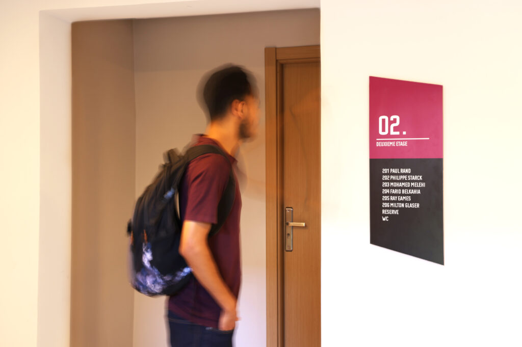 Un étudiant avec un sac à dos se tient devant une porte, à côté d'un panneau indiquant le deuxième étage à l'école de design