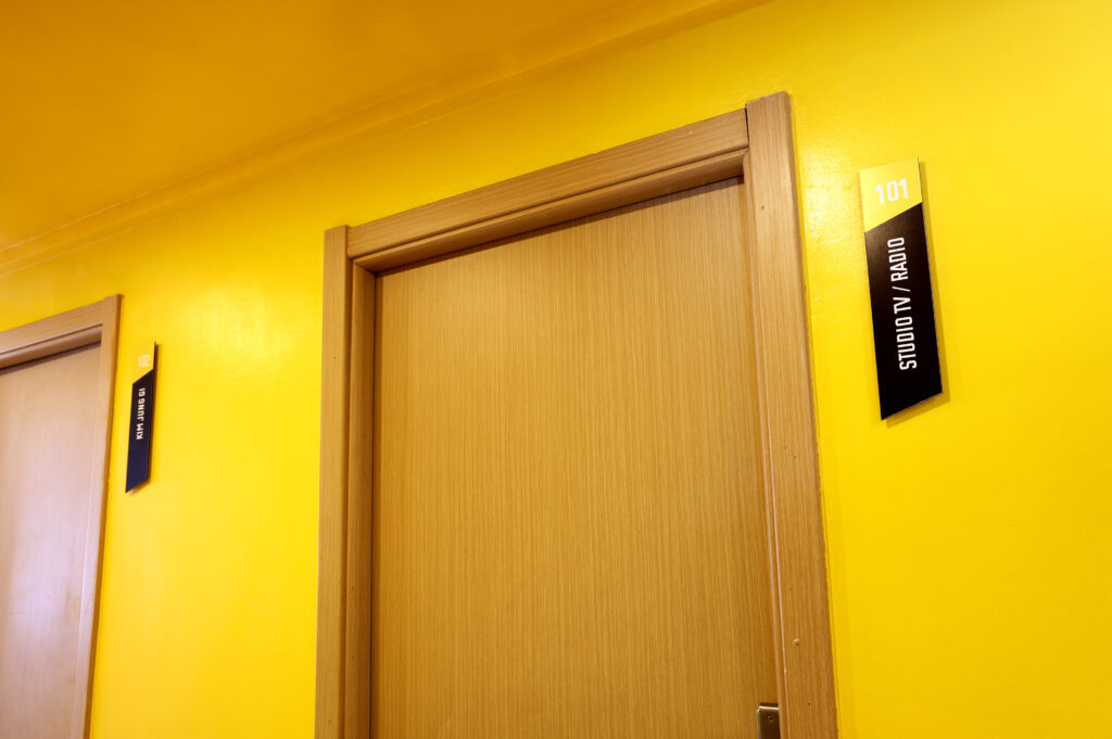 Porte en bois avec un panneau indiquant le studio TV/Radio à l'école de design sur un mur jaune