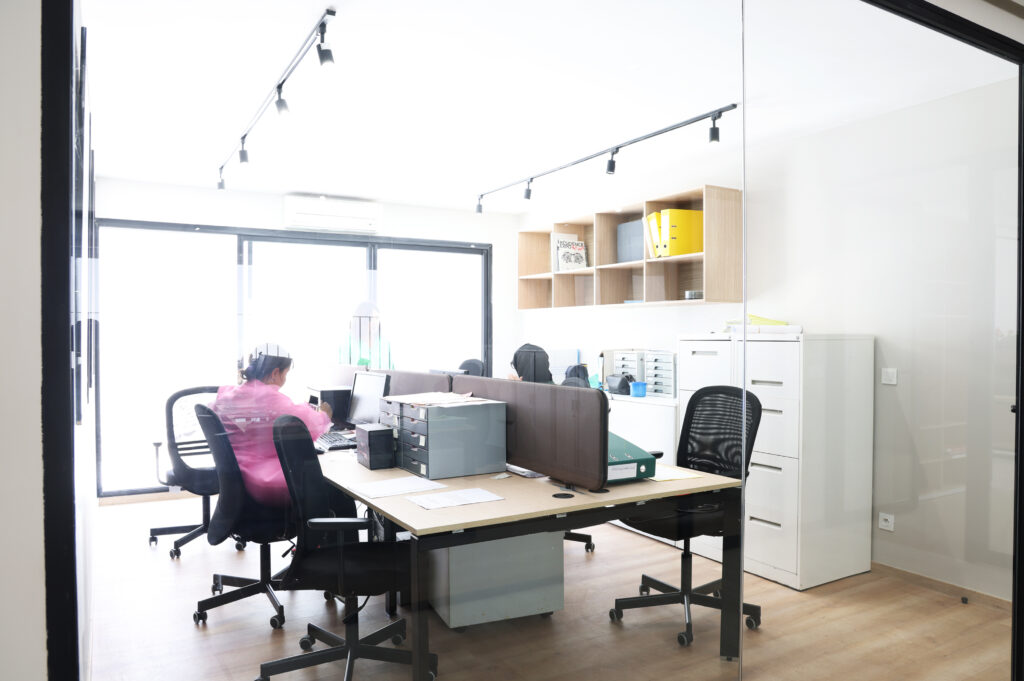 Vue d'un bureau moderne avec une employée travaillant sur son ordinateur. Des étagères au mur contiennent des classeurs et des fournitures de bureau, tandis qu'un environnement lumineux favorise la productivité