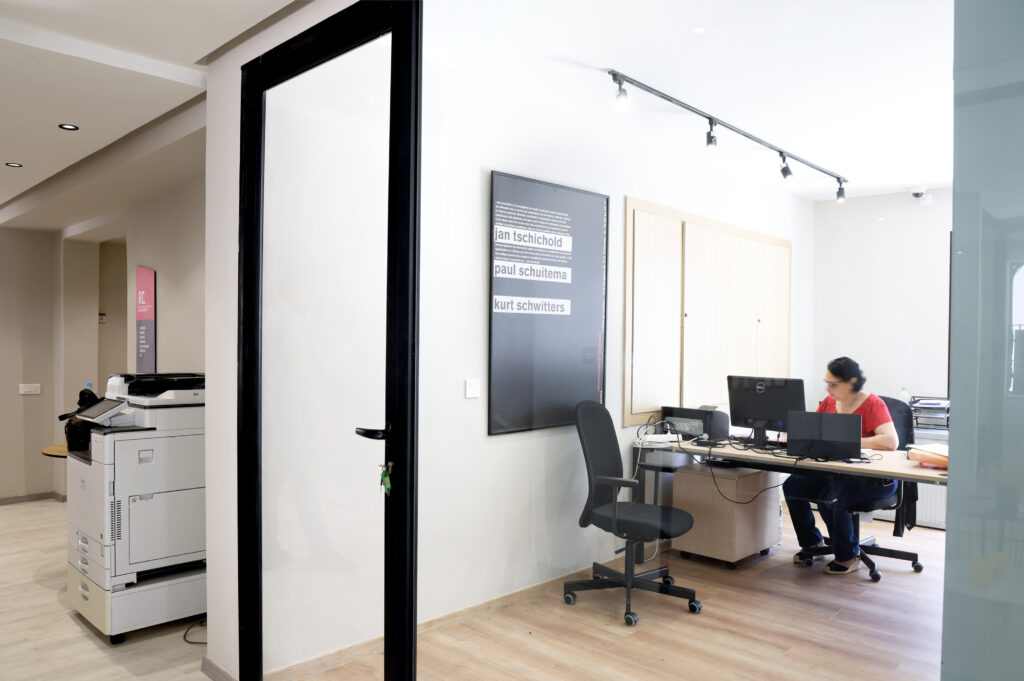 Vue d'un bureau moderne où une employée travaille à son ordinateur. À gauche, une imprimante est visible, et des affiches sont accrochées au mur, ajoutant une touche artistique à l'espace