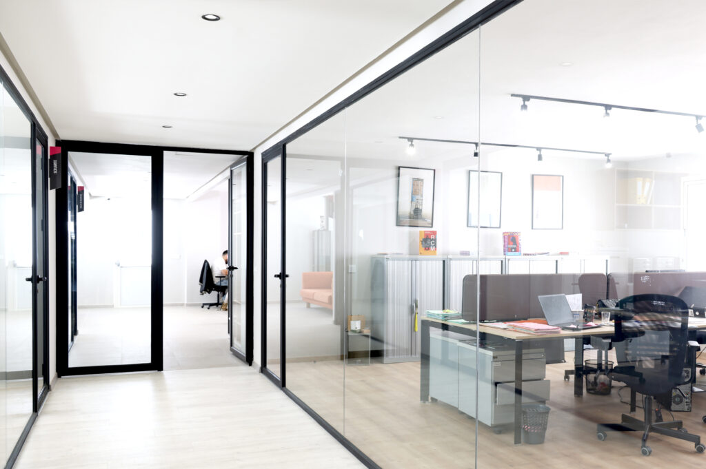 Vue d'un couloir lumineux dans un bureau moderne avec des espaces de travail vitrés. On aperçoit des employés au travail derrière des cloisons en verre, créant une atmosphère collaborative et dynamique