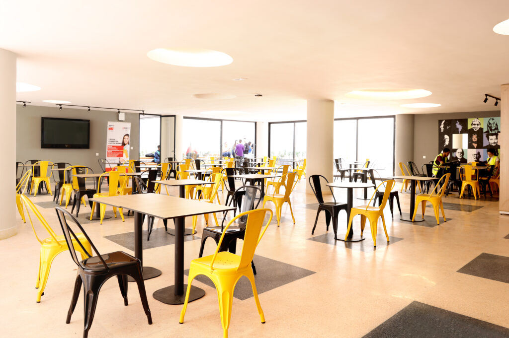 Vue d'un espace de restauration moderne avec des tables et des chaises jaunes et noires, un écran de télévision en arrière-plan, et des fenêtres offrant une vue sur l'extérieur