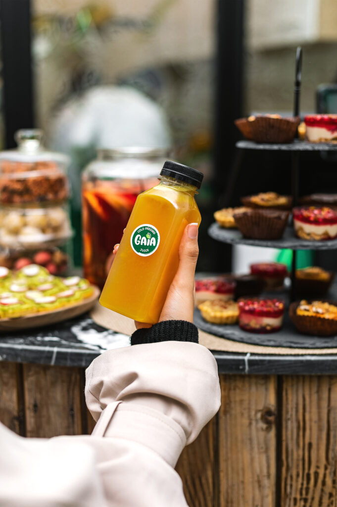 Personne tenant une bouteille de jus pressé à froid GAIA Juice devant un assortiment de desserts.