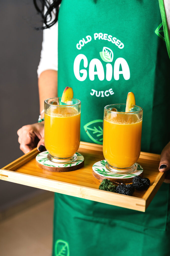 Serveuse de GAIA Juice Lab avec tablier vert, tenant un plateau de jus pressés à froid avec des tranches de fruits