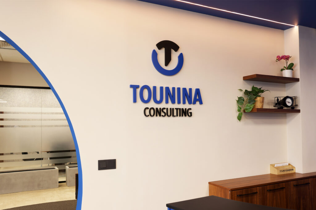 Vue rapprochée du logo de Tounina Consulting sur un mur décoratif, avec des étagères contenant des plantes et une horloge moderne en arrière-plan