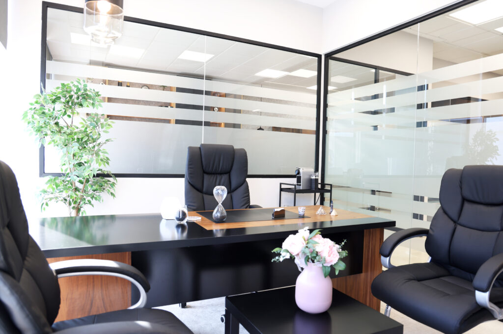 Vue d'un bureau avec une chaise en cuir noir, un bureau noir, des accessoires de bureau, une plante verte et des parois en verre dépoli, offrant une ambiance professionnelle et accueillante