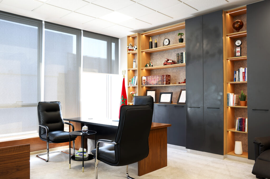 Vue d'un bureau contemporain avec un bureau noir, des chaises en cuir, des étagères en bois illuminées et des éléments de décoration marocaine, dont un drapeau et des diplômes