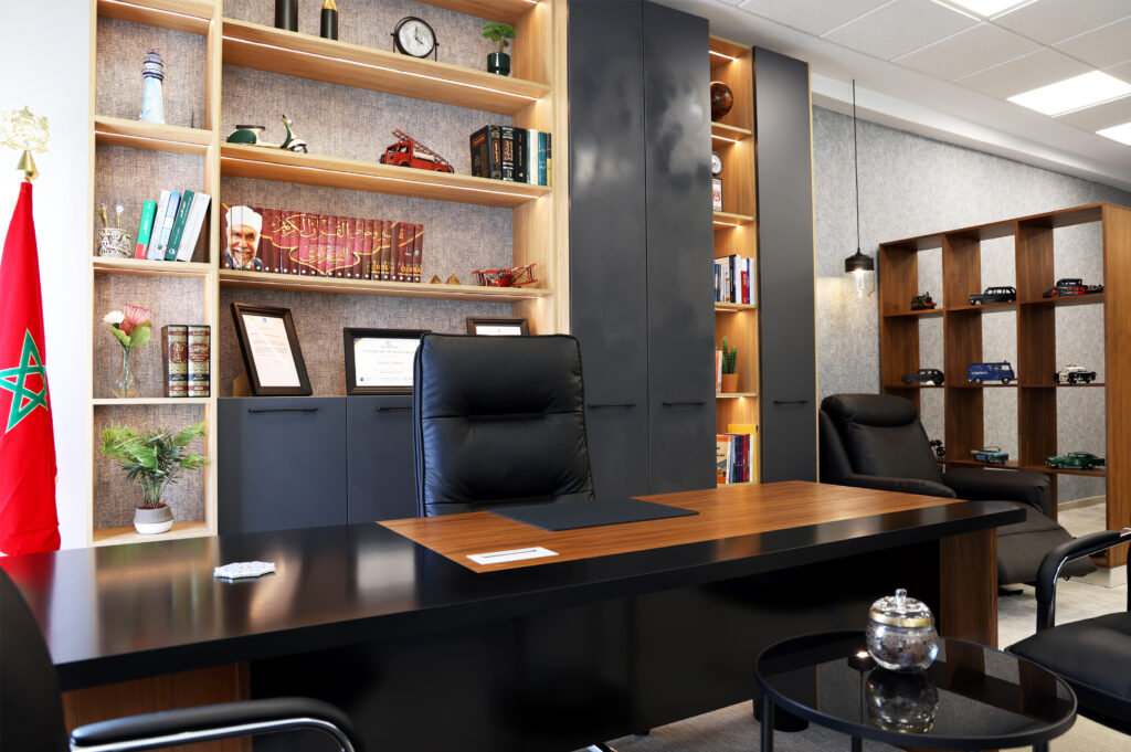 Vue d'un bureau moderne avec un grand bureau noir, des étagères en bois éclairées contenant des livres et des objets de décoration, un drapeau marocain, et un coin confortable avec un fauteuil en cuir