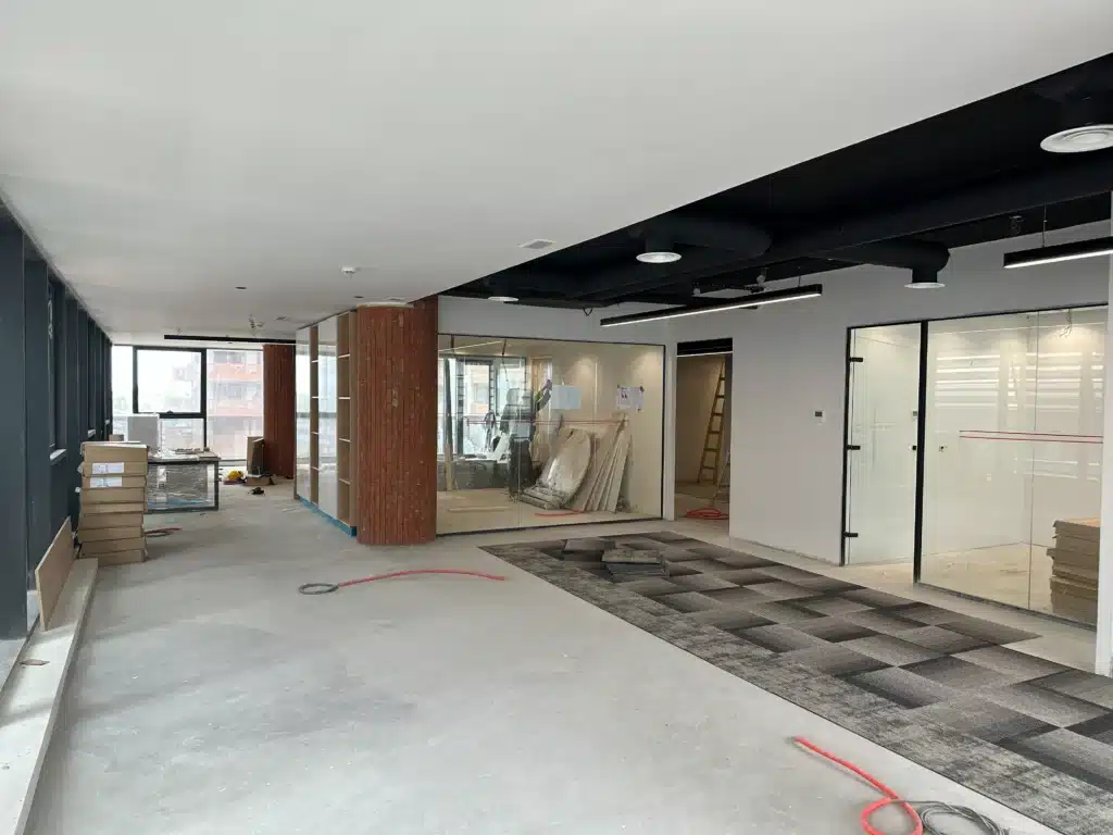 Chantier de bureaux en phase de finition avec poteaux habillés de briques rouges, cloisons vitrées et pose de moquette.