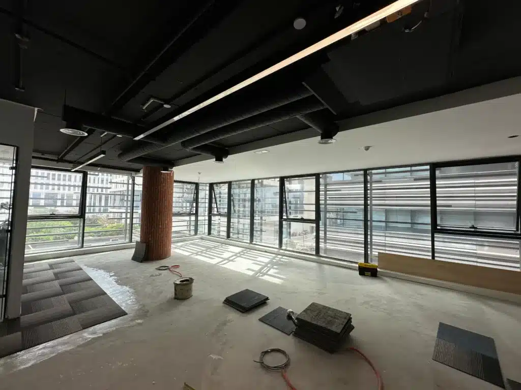 Vue d'ensemble d'un bureau en travaux montrant la pose de dalles de moquette et l'installation de luminaires LED suspendus.